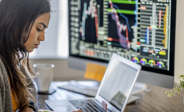 Assise à un bureau, une femme travaille sur son ordinateur portable, avec un moniteur affichant des données financières à l’arrière-plan.