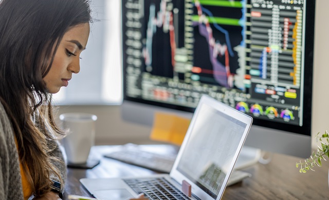 Woman sitting at desk in front of laptop and a monitor displaying financial charts