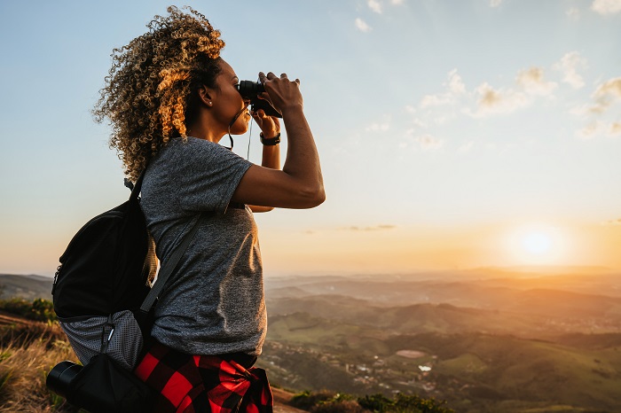 Une femme en randonnée pédestre au sommet d'une montagne, jumelles en main et regardant la vue au coucher du soleil.